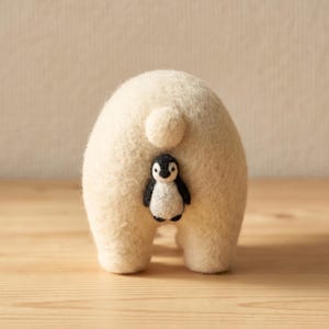 Op de afbeelding: Een naaldgevilte, crèmekleurige ijsbeerfiguur van achteren gezien, met een kleine ronde staart en een piepklein zwart-wit pinguïnfiguurtje bevestigd in het midden van de rug van de beer, gemaakt met een zachte, getextureerde wolvezelafwerking.