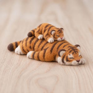 Op de afbeelding: Een sculptuur van naaldviltwol van een grote tijger die ligt met een kleinere tijgerwelp die op zijn rug rust. Beide tijgers hebben een oranje vacht met donkerbruine strepen, witte buiken en gesloten ogen, alsof ze slapen.