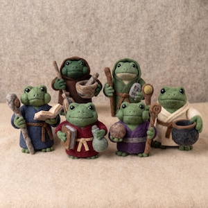 May include: Six needle-felted frog figurines dressed in robes. One wears a brown robe with a broom, one green with a stone, one blue with a staff and book, one red with a book and potion, one purple with a staff and orb, and one cream with a cauldron.