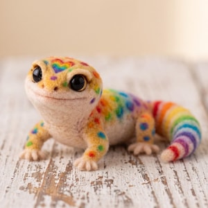 May include: A needle-felted leopard gecko figurine with a cream-colored body, featuring a rainbow-colored heart on its head, multicolored spots along its back, and a vibrant rainbow-striped tail. The gecko has large, glossy black eyes.