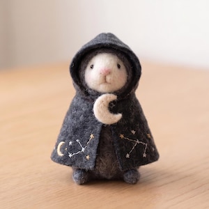 Puede incluir: Una pequeña figura de animal de fieltro con cara blanca y cuerpo gris oscuro, que lleva una capa con capucha gris oscuro. La capa presenta estrellas, constelaciones y una luna creciente bordadas en oro y blanco, con un detalle de luna de fieltro crema.