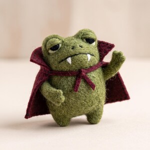 May include: A needle-felted green frog figurine wearing a dark maroon cape with a high collar. The frog has two white fangs protruding from its mouth, dark eyes, and one arm raised in a waving gesture. The texture of the felt is visible throughout.
