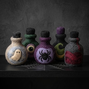 Puede incluir: Cinco botellas de poción pequeñas y redondas de lana fieltrada con tapones de corcho negro. Las botellas presentan diseños de fieltro con aguja que incluyen un fantasma blanco, un ojo verde, una araña negra, una luna creciente amarilla y un patrón de remolinos rojos sobre una base oscura.