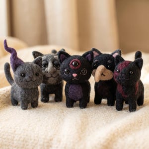 Può includere: Una collezione di cinque statuette di gatti in lana infeltrita. I gatti presentano vari design, tra cui uno con una coda viola, uno con un volto umano, uno con un terzo occhio sulla fronte, uno con un naso a becco e uno con segni rossi.