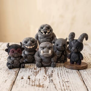 May include: A collection of six needle-felted figures in various shades of gray and black. The figures include creatures with horns, tusks, and wide grins, all crafted with a textured wool finish. One figure stands on a small circular wooden base.