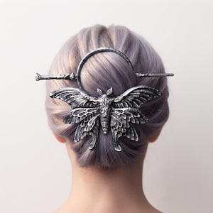 Puede incluir: Un accesorio para el cabello de tono plateado con un diseño detallado de polilla con alas texturizadas y un cuerpo central. La polilla está unida a un marco circular y a un pasador recto que asegura el cabello en un moño decorativo.