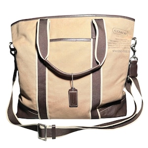 May include: A tan canvas tote bag with dark brown leather trim and dark brown fabric stripes. It features a detachable shoulder strap, two top handles, and a hanging leather luggage tag. The front displays a Coach logo and text patch.