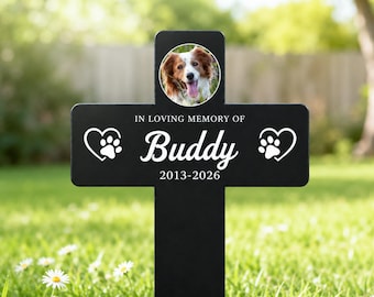 Op de afbeelding: Een zwarte kruisvormige gedenkpaal voor in de tuin met een ronde foto van een bruin-witte hond aan de bovenkant. De tekst luidt IN LOVING MEMORY OF Buddy 2013-2026, geflankeerd door twee witte harticonen met pootafdrukken aan weerszijden.