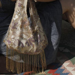 May include: A cream-coloured hobo bag featuring intricate floral embroidery in shades of purple and gold. The surface is embellished with shimmering sequins and beads, finished with a long, dangling gold fringe along the bottom edge.