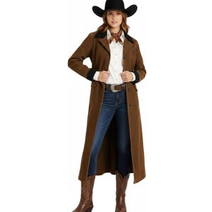 May include: A long brown duster coat worn over a white button-down shirt and blue jeans. The outfit includes a black cowboy hat, a brown neck scarf, a brown leather belt with a silver buckle, and brown leather cowboy boots with decorative stitching.