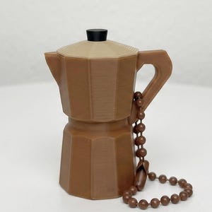 Op de afbeelding: Een bruine moka-pot sleutelhanger met een achthoekig lichaam, een lichtbruin deksel met een kleine zwarte knop en een bruine kralenketting bevestigd aan het handvat. Het object heeft een gladde, matte afwerking en een geometrische vorm.