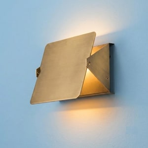 May include: A wall-mounted light fixture with a brushed brass finish, featuring a flat, rectangular metal plate angled outward. The design includes triangular side panels secured with visible screws, emitting a warm glow from the interior.