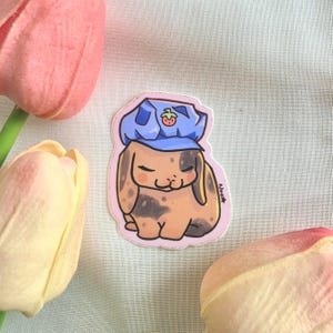 May include: A die-cut sticker featuring a tan and brown lop-eared rabbit with closed eyes, wearing a blue cap adorned with a small red strawberry icon. The illustration has a light pink border and includes the signature Neo star on the right side.