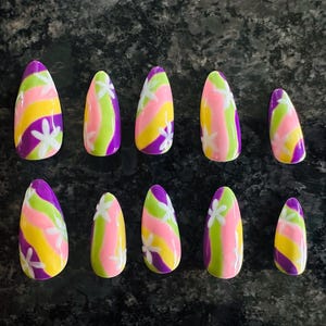 May include: A set of ten almond-shaped press-on nails featuring a vibrant wavy pattern in shades of purple, lime green, yellow, and pink. Each nail is decorated with small, white, five-petaled flower motifs scattered across the colorful stripes.