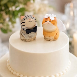 May include: Two small cat figurines placed on top of a two-tiered white cake. One cat is grey with black stripes and wears a black bow tie. The other cat is orange with a white bow and a white veil attached to its head. Both cats have closed eyes.