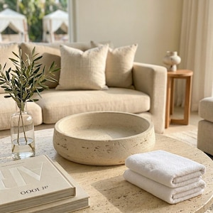 May include: A round, beige, porous stone bowl sits on a matching stone coffee table next to a clear glass vase containing olive branches, two stacked white folded towels, and a beige book with the text at 1000 on the cover.
