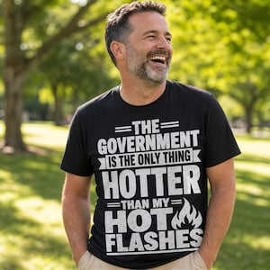 Puede incluir: Una camiseta negra de manga corta con texto blanco desgastado que dice: THE GOVERNMENT IS THE ONLY THING HOTTER THAN MY HOT FLASHES, junto a un pequeño gráfico de una llama al lado de la palabra HOT.