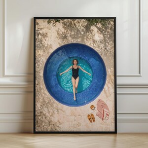 May include: A framed wall art print featuring an aerial view of a person in a black swimsuit floating in a circular blue tiled pool. Beside the pool, a pair of sandals and a red and white striped towel rest on a light-colored stone patio surface.