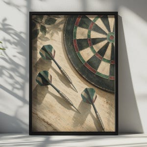 May include: A framed wall art print featuring a circular dartboard with black, red, and green segments alongside three darts with matching green, red, and white flight patterns. The composition is set against a light, textured wooden surface.