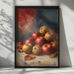 May include: A framed still life print featuring a cluster of red and yellow apples resting on a light-coloured wooden surface. Scattered dried autumn leaves surround the fruit, set against a dark, moody background with blended red and blue tones.