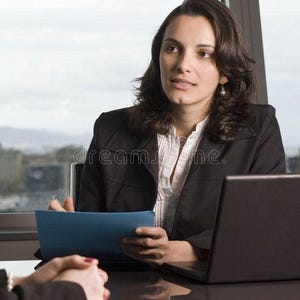 May include: A professional wearing a black blazer over a white button-down shirt, holding a blue folder while seated at a desk with a black laptop. The individual has dark hair and is positioned in an office setting with a large window view.