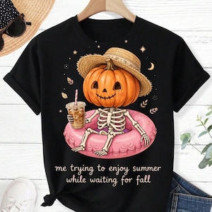 May include: A black t-shirt featuring a graphic of a skeleton with a pumpkin head wearing a straw hat, sitting in a pink inflatable pool ring while holding an iced coffee. The text reads: me trying to enjoy summer while waiting for fall.