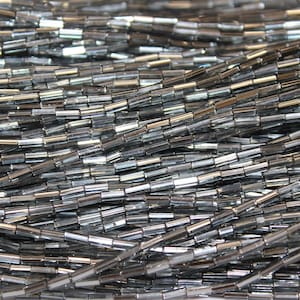 May include: A collection of metallic gunmetal gray bugle beads arranged in multiple parallel strands. Each individual bead features a cylindrical, elongated shape with a reflective, polished surface that catches light, creating a shimmering effect.