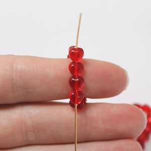 May include: A series of small, translucent red heart-shaped beads strung onto a thin, straight gold-colored wire. The beads are uniform in size and shape, featuring a smooth, glossy finish that catches the light, creating a vibrant red color.
