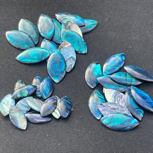 May include: A collection of marquise-shaped abalone shell cabochons featuring iridescent surfaces with vibrant blue, green, and purple hues. These polished shell pieces display natural swirling patterns and a high-gloss finish, arranged in three distinct clusters.