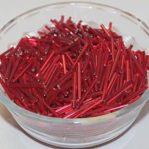 May include: A clear glass bowl filled with a large quantity of small, cylindrical, bright red glass bugle beads. The beads have a smooth, reflective surface and are uniform in shape, creating a dense pile of vibrant red within the transparent container.