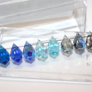 May include: A row of eight faceted, teardrop-shaped crystal beads in a gradient of blue and grey tones, ranging from deep cobalt to translucent light blue and dark charcoal, threaded on a thin silver wire inside a clear plastic storage container.