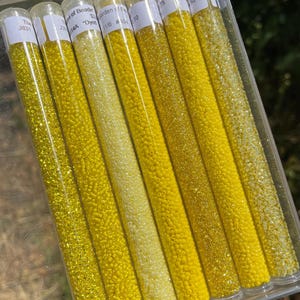 May include: Seven clear plastic tubes filled with small yellow seed beads in varying shades and finishes, ranging from pale lemon to deep golden yellow. Each tube features a white adhesive label with printed text at the top, organized in a clear container.