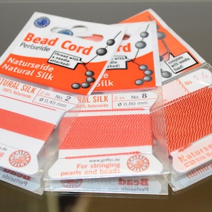 May include: Three cards of orange natural silk bead cord for stringing pearls and beads. The packaging includes text such as Bead Cord, Natural Silk, 2m, and specific diameters of 0.45mm and 0.80mm. Each card features a needle attached to the cord.