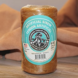 May include: A cylindrical spool of natural-colored artificial sinew wrapped in clear plastic. The teal label features the text Artificial Sinew, Tendon Artificiel, Gudebrod Thread Company, Established 1870, 8 oz, 300 yds, 900 ft, 70 lb, 5 ply, and a barcode.