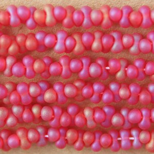 May include: A collection of pink, hourglass-shaped beads strung together in multiple horizontal rows. Each bead features a smooth, matte finish with subtle iridescent highlights, creating a uniform, repeating pattern across the entire display.