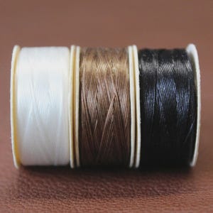 May include: Three small spools of thread arranged side by side, featuring one white spool, one brown spool, and one black spool. Each spool has a light-colored cardboard core and is tightly wound with a smooth, braided synthetic cord material.