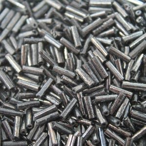 May include: A collection of small, cylindrical, metallic dark gray bugle beads. Each bead features a smooth, reflective surface with a hollow center, arranged in a dense, overlapping pile that creates a textured, monochromatic appearance.