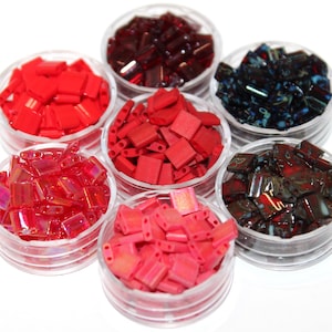 May include: A collection of seven clear circular containers filled with small, rectangular, two-hole beads in various colours including bright red, matte red, dark burgundy, iridescent pink, and dark blue with multi-coloured metallic flecks.