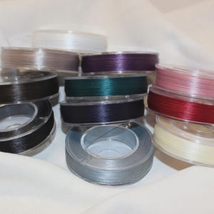 May include: A collection of circular plastic spools wound with thin, colorful nylon cord. The assortment includes spools in shades of white, beige, dark brown, black, deep purple, forest green, light pink, dark red, cream, and metallic silver.