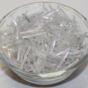 May include: A clear glass bowl filled with a large quantity of long, thin, cylindrical clear glass bugle beads. The beads have a subtle iridescent finish that reflects light, creating a shimmering effect throughout the collection of craft supplies.