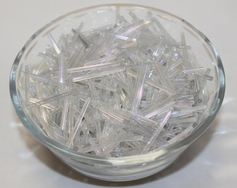 May include: A clear glass bowl filled with a large quantity of long, thin, cylindrical clear glass bugle beads. The beads have a subtle iridescent finish that reflects light, creating a shimmering effect throughout the collection of craft supplies.