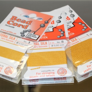 May include: Three cards of yellow Griffin natural silk bead cord in clear plastic packaging. Each card features the text Bead Cord, Natural Silk, 100% Naturseide, and For stringing pearls and beads. The cards show diameters of 0.50 mm and 0.80 mm.