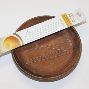May include: A set of five silver-colored curved needles in a white and yellow cardboard package labeled Basic Elements and Spin & String, resting inside a round, dark brown wooden bowl with a smooth, polished finish and visible grain.