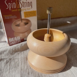 May include: A light-colored wooden bead spinner tool with a central metal spindle and a knurled top knob. The tool is positioned in front of its original product packaging, which features the text Spin & String bead stringing tool and The Bead Smith.