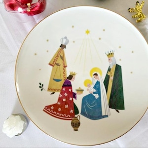 Może przedstawiać: Okrągły biały talerz ceramiczny ze złotym obrzeżem, ozdobiony kolorową ilustracją sceny Bożego Narodzenia. Wzór przedstawia Świętą Rodzinę oraz trzy postacie w szatach, z żółtą gwiazdą powyżej i małymi złotymi gwiazdkami rozrzuconymi wokół.