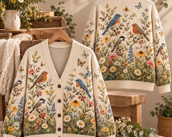 May include: A cream-colored knit cardigan and a matching pullover sweater, both featuring a detailed pattern of various colorful songbirds, butterflies, and wildflowers including daisies and lupines, with a button-front closure on the cardigan.