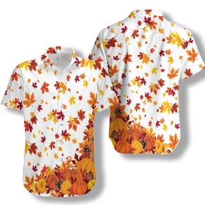 May include: A short-sleeved button-down shirt featuring a white background with a scattered pattern of autumn leaves in shades of red, orange, and yellow. The bottom hem area displays a cluster of orange pumpkins and pinecones.