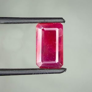 May include: A vibrant, deep red gemstone cut into a rectangular emerald shape. The stone features faceted edges that reflect light, creating a clear, polished appearance. It is held securely between the two black-tipped ends of metal tweezers.