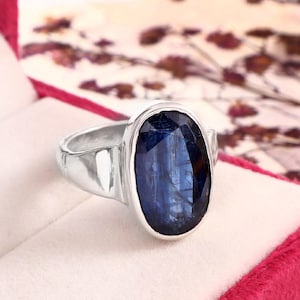 May include: A silver ring featuring a large, oval-shaped, faceted deep blue kyanite gemstone set in a smooth metal bezel. The band is polished silver with a tapered design, showcasing the dark blue stone's natural inclusions and reflective surface.