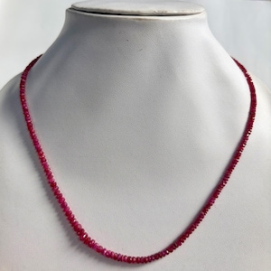 May include: A single-strand necklace composed of numerous small, faceted, deep red ruby beads. The beads are uniform in size and shape, strung together to form a continuous circular chain that rests evenly around the neck display form.
